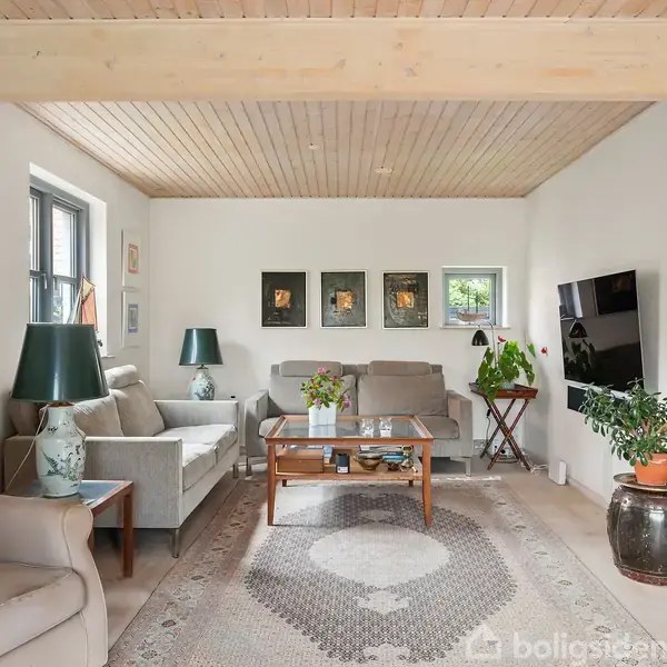 Stue med to beige sofaer og et sofabord, dekoreret med planter og lamper. Væggene har kunstværker, og et fladskærms-tv er monteret på højre væg. En plakat hænger til venstre.
