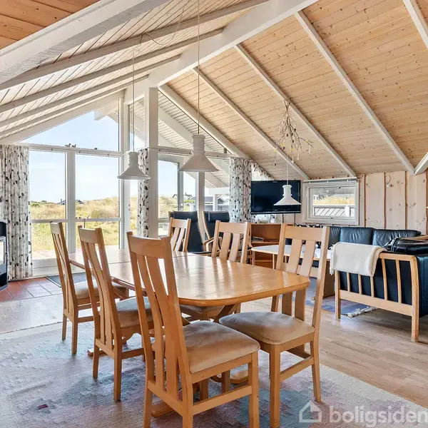 Spisebord med seks træstole står i et lyst rum. Rummet har skråloft, store vinduer med udsigt til natur, og indeholder også en sofa og brændeovn i baggrunden.
