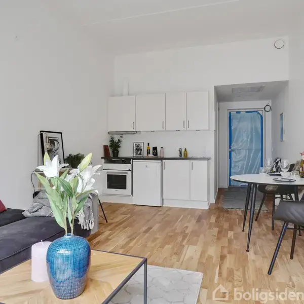 Stue med sofa, sofabord og vase med blomster; tekøkken i baggrunden med hvidt skab; rundt spisebord med to stole. Lysindfald gennem venstre væg.