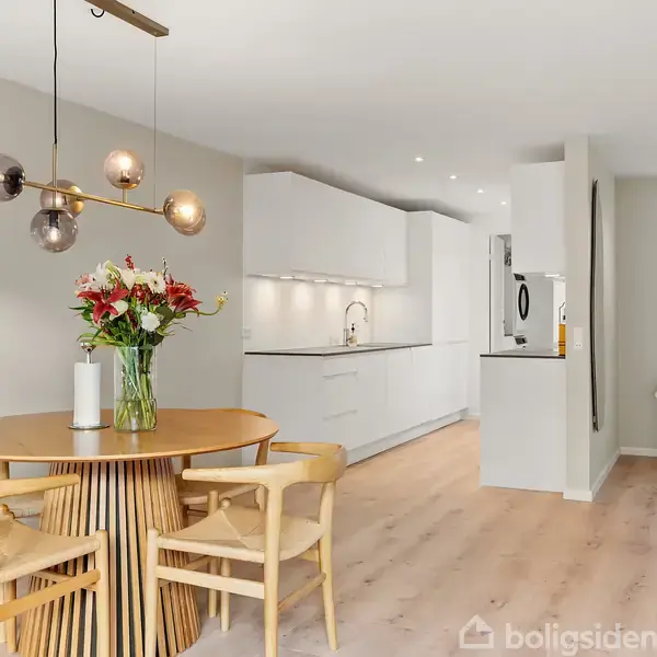 Et rundt spisebord prydet med blomster står i en moderne stue/køkken. Der er hvide skabe, en ovn indbygget i væggen og træstole omkring bordet.