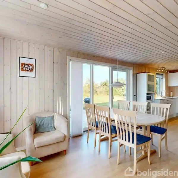 Stue og køkken i åben plan med trævægge og loft. En sofa står ved glasskydedøre til udendørs areal, og et spisebord med stole er centralt. Køkkenet har hvide skabe.