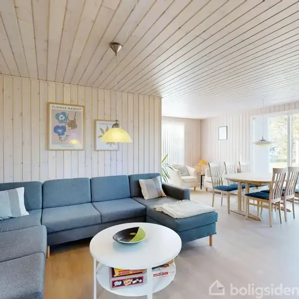 En stor stue med en blå hjørnesofa fyldt med puder centreret om et rundt sofabord. I baggrunden er der et spisebord og store vinduer, der slipper lys ind.