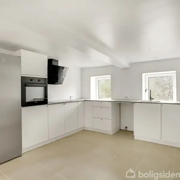 Et moderne køkken, domineret af hvide skabe og en grå kølefryseskab, står i et lyst rum med vinduer, hvilket skaber et minimalistisk og funktionelt miljø.