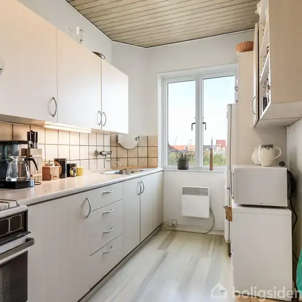 Et lyst køkken med en ovn og kogeplader til venstre, skabe langs væggen, og en vask nær et vindue. En elkedel, brødrister og forskellige krydderier er synlige på bordpladen.