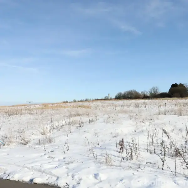 Sneklædt mark ligger stille under en klar blå himmel; i forgrunden ses et vejskilt med tekst. Træer danner en horisontlinje i baggrunden.