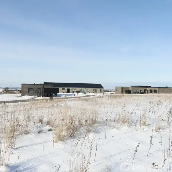 Sneklædte moderne huse står i en åben mark. Sne dækker jorden, mens en blå himmel strækker sig over området. Huse er spredt med mellemrum og omgivet af åbne arealer.