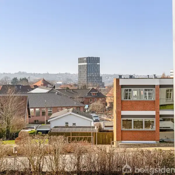 Et bylandskab med flere bygninger, herunder et højt, moderne tårn i baggrunden. Forgrunden har boligblokke og hække, set fra et vindue. Klar, blå himmel uden tekst.