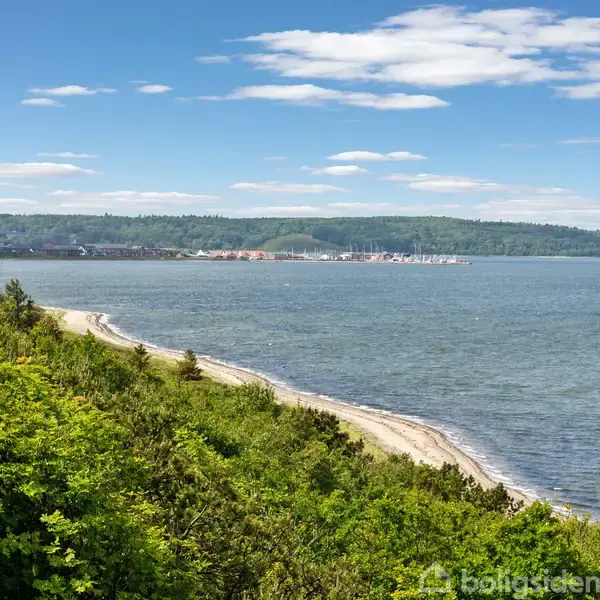 Kystlinje med sandstrand strækker sig langs en frodig, grøn skov. Havet er roligt under en klar himmel med spredte skyer. I det fjerne ses en lille havneby.