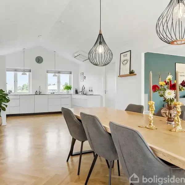 Langt træbord med grå stole står i en spisestue. Hængelamper over bordet kaster lys ned. I baggrunden ses et lyst køkken med store plantekrukker og moderne indretning.