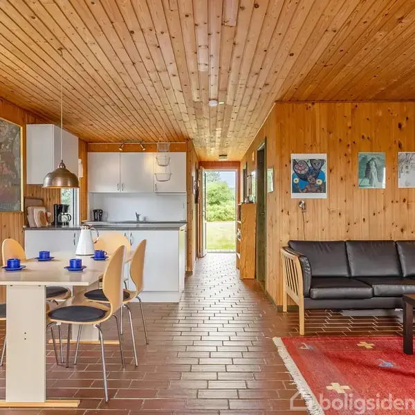 Spisebord med blå kopper står ved siden af sort sofa. Køkken i baggrunden, træpaneler på vægge og loft, rød tæppe og billeder på væggen. Udsigt til grøn have.