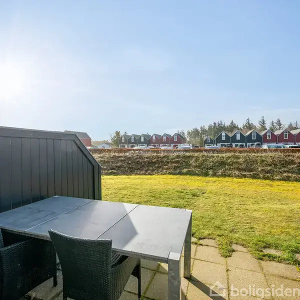 Bord med to stole står på terrasse; solen skinner; i baggrunden ses grøn græsplæne og række røde og blå rækkehuse.