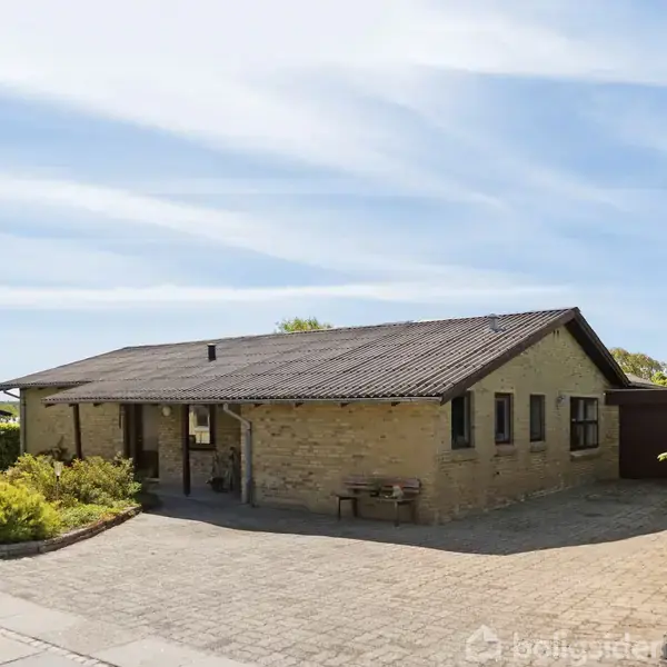 Et enfamiliehus står stille i solen, omgivet af en velholdt flisebelagt indkørsel og grønne buske, under en blå himmel med lette skyer.