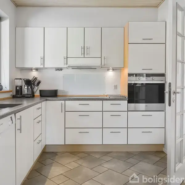 Et lyst køkken med hvide skabe og skuffer monteret på væggen. Køkkenet indeholder en ovn, opvaskemaskine og en vask med udsigt til en have gennem et vindue.