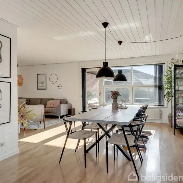 Et spisebord med sorte stole står centralt i et lyst rum. Sort belysning hænger over bordet. I baggrunden ses en sofa, kunst på væggen og planter.