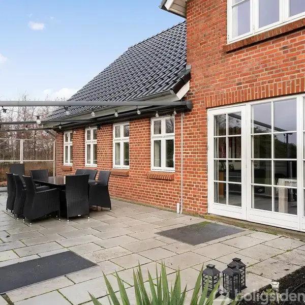 Rødt murstenshus med sort tag, store vinduer og gårdsplads. Havemøbler under glastag på terrasse i moderne have. Ingen tekst er synlig.