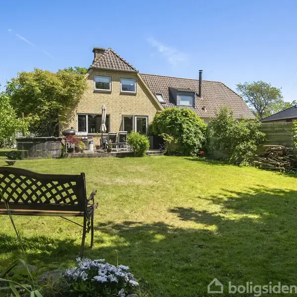 Bænk står stille i en velplejet græsplæne med blomster i forgrunden. I baggrunden ses et gult murstenshus med to etager, omgivet af buske og træer.