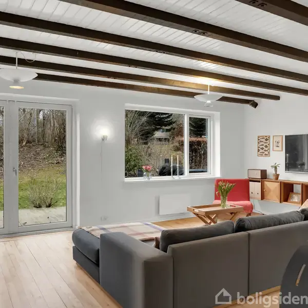 Grå sofa står i en stue med trægulv og lofter med træbjælker. Store vinduer og glasdøre giver udsigt til en grøn have. En pejs er synlig til højre.
