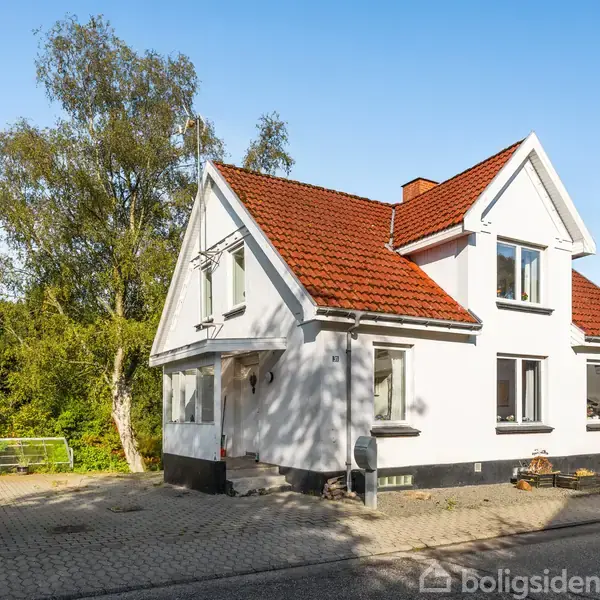 Hvidt hus med rødt tag står på en brostensbelagt grund. Omkring huset er der træer og anden beplantning. Solskin skaber skygger på facaden.