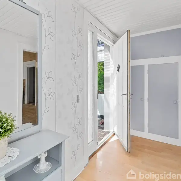 Et skab står i en lys entré med en åben dør, der leder udenfor. En plante er placeret på et lille bord under et spejl. Veggene har blomstermønster.