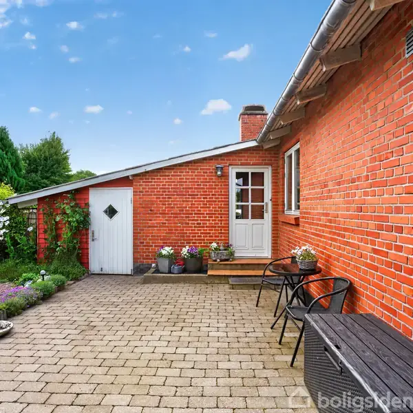 Rødt murstenshus står ved siden af en stenbelagt gårdhave med to sorte stole og et bord. Omkring er der velplejede haver med grønne planter og farverige blomster.