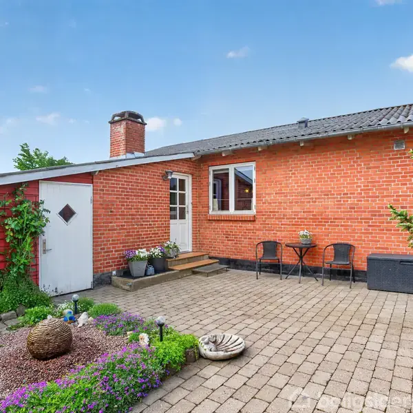 Et rødt murstenshus med en hvid dør åbner ud til en flisebelagt terrasse. To stole og et bord står ved siden af døren. Haven er dekoreret med blomster og buske.