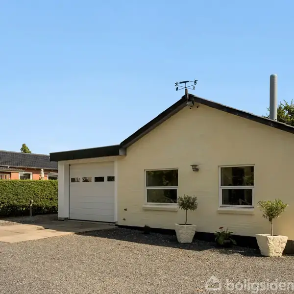 Et hvidt, enkelt-etagers hus med fladt tag står stille på en grusbelagt gårdsplads. To buske i krukker flankerer hovedvinduet. Grøn hæk omkranser ejendommen.