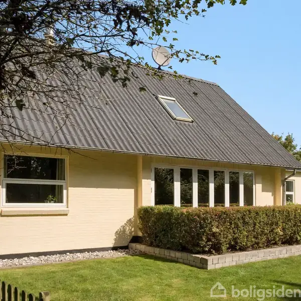 Et gult murstenshus med skråt tag og vinduesfacade står i en grøn have med hæk og stakit. Et skur ses i baggrunden.