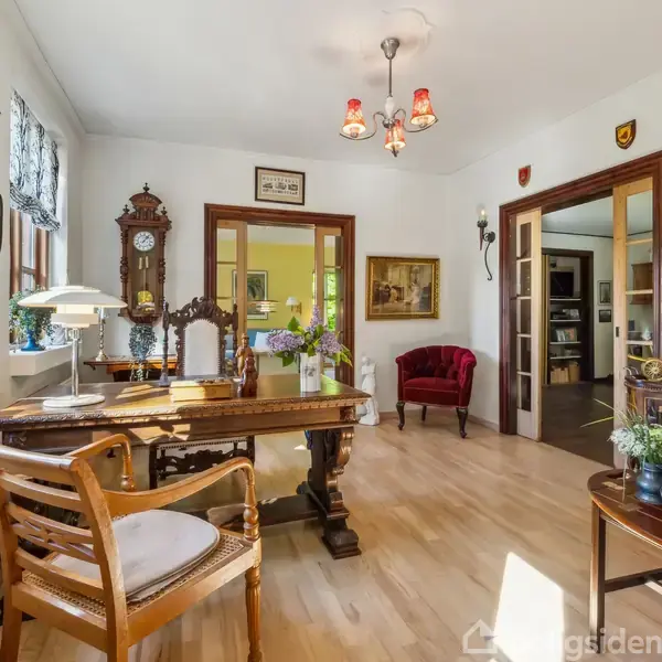 Kontor med træbord og -stol ved vindue, omgivet af antik indretning som en væg-ur og malerier. Dobbelt døre fører til en lys stue med røde møbler.