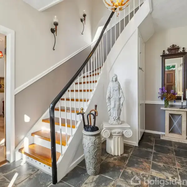 Trappe drejer opad i entre, keramikkrukke med paraplyer ved siden af statue. Spejl og blomsterarrangement står på konsolbord. Åben dør viser spisestue med møbler.