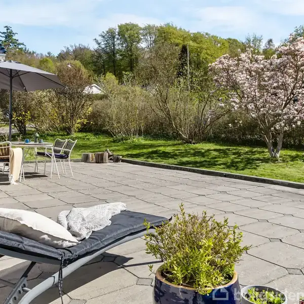 Liggestol med puder står på flisebelagt terrasse ved siden af en parasol og et bord med stole. En blomstrende træ omkranser græsplæne i en have.