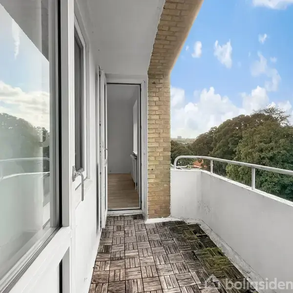 En balkon med flisebelægning fører ind til en åben dør. Udsigt til træer og en blå himmel med skyer. Væggene er hvidmalede med en murstensbue.