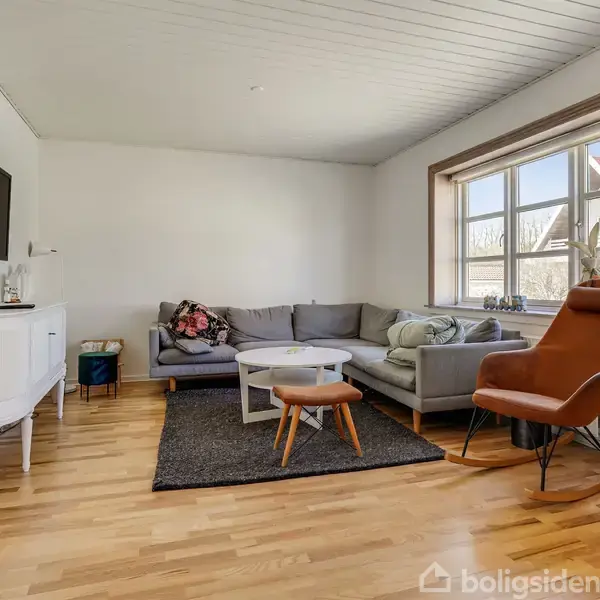 Sofa står i hjørnet med gråt betræk, ved siden af et hvidt sofabord og brun lænestol. Fladskærms-tv hænger på væggen; stort vindue lader lys ind i stuen.