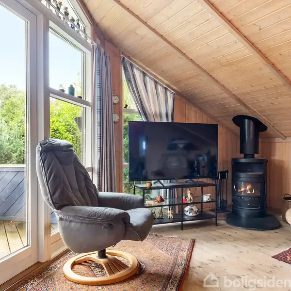 En lænestol står ved en stor glasdør indrammet af gardiner, der fører ud til en terrasse. Ved siden af er en brændeovn og et tv i en træbeklædt stue.
