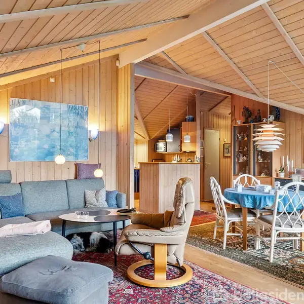 Stue med blå sofa og træstol er placeret omkring et rundt bord. Langbord med blå dug ses ved spiseplads. Træpaneler og loft pryder rummet sammen med vægkunst.