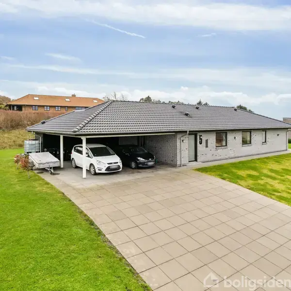 Et moderne hus af mursten med skråt tag står på en velplejet græsplæne. To biler er parkeret i en carport ved siden af huset. Skyer danner en lys blå himmel i baggrunden.