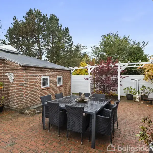 Terrassemøbler står omkring et spisebord på en brolagt gårdhave. Omgivet af en murstensbygning og en hvid hegnspergola med flere planter i krukker, er der frodige træer i baggrunden.
