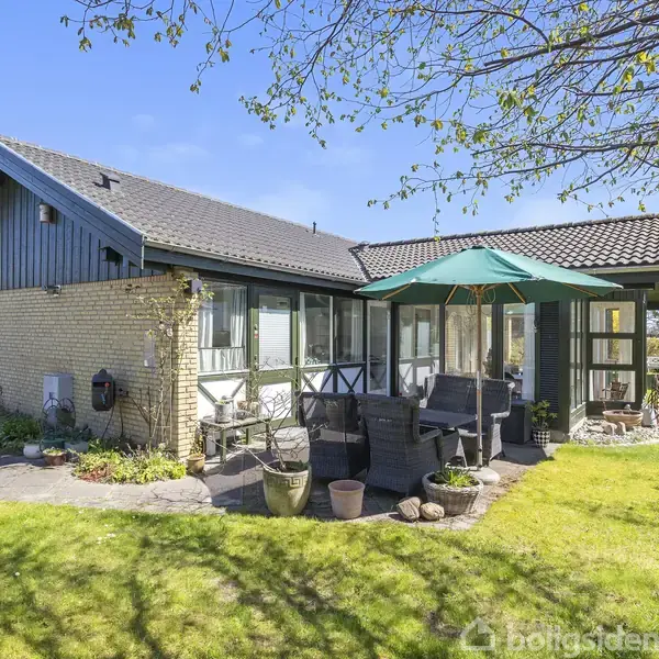 Hus med fladt tag i gule mursten, havemøbler under parasol i solen, omgivet af grøn have med træer og terrasse.