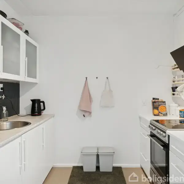 Et kompakt køkken med hvide skabe og apparater. Til venstre står en elkedel ved siden af en vask. Til højre er en ovn med gryder, pander og morgenmadsprodukter ovenpå. På bagvæggen hænger to håndklæder og to skraldespande står nedenunder.