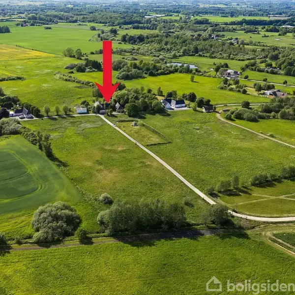 Et stort grønt landskab med spredte gårde. En rød pil peger mod et hus blandt træer. Marker omgiver området, og veje ses gennem landskabet. Ingen tekst bortset fra "boligsiden" vandmærke.