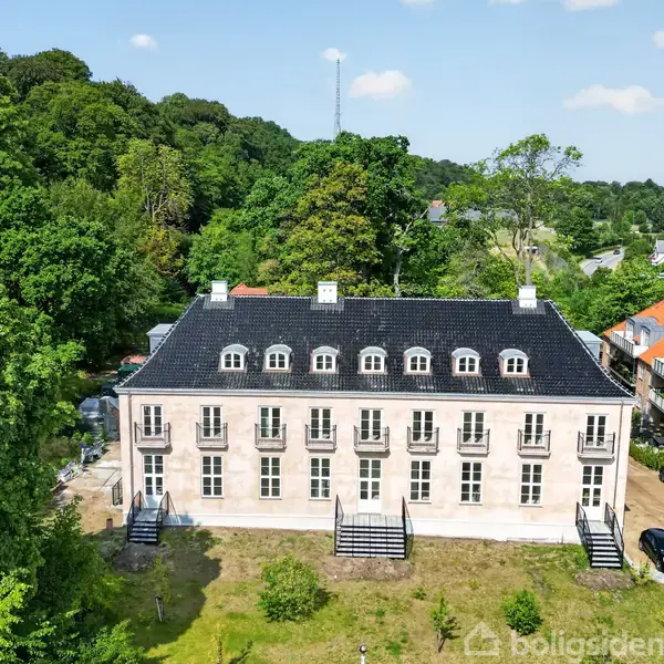 Stort, hvidt hus med sort tag og hvælvede vinduer, omgivet af grønne træer og et åbent område. Baggrunden viser omkringliggende huse i en skovklædt landskab.