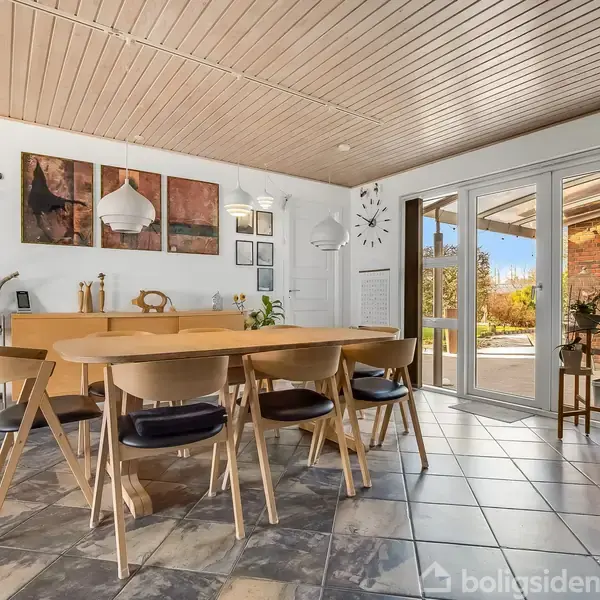 En spisebordgruppe med træstole, der står på et flisegulv. Store vinduer giver udsigt til en terrasse med planter udenfor. Kunst hænger på væggen, og en trappe ses til højre.