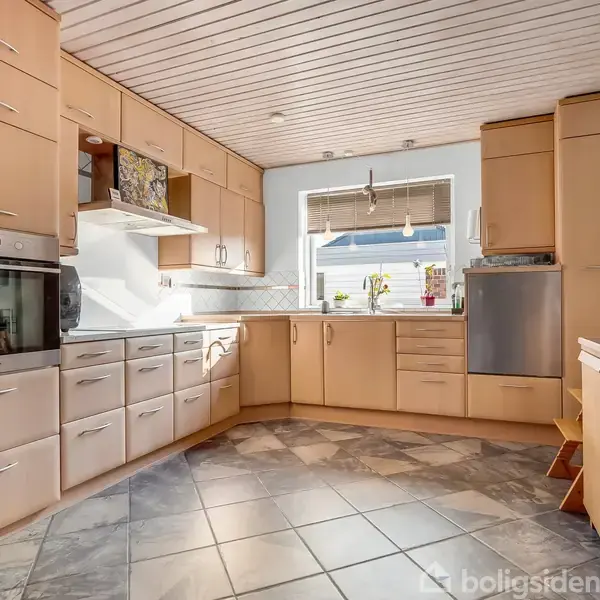 Et stort køkken med træskabslåger og hvidevarer. Vindue med lysindfald omgivet af flisegulv og lyse vægge, kaffemaskine på en bordplade. Skuffer og skabe fylder væggene.