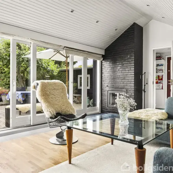 Stolen domineret af en glasbord og en lænestol med fåreskind. Rummet har stor glasåbning mod en terrasse med grøn udsigt, og en sort pejs ved hvidmalede vægge.