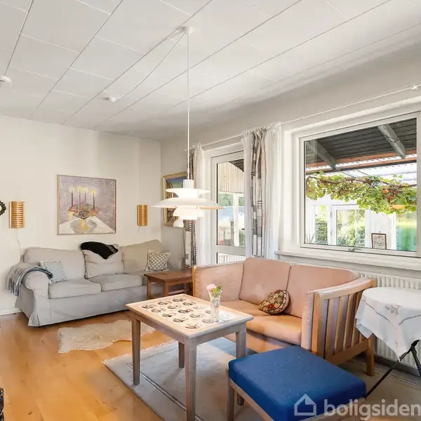 En stue med en grå sofa og et lille træbord i midten. Rummet er lyst med store vinduer, lyse træmøbler og en hængende lampe. Gardiner hænger ved vinduerne.