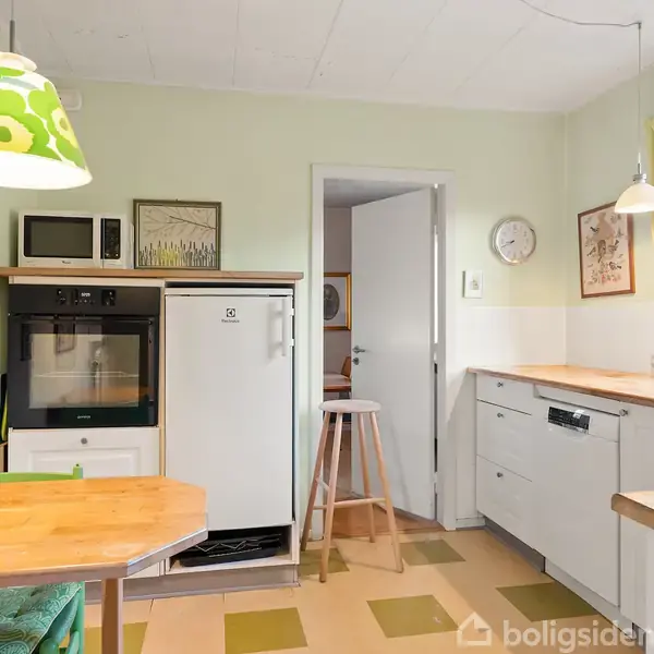 Et køkken med opvaskemaskine til højre, et køleskab og ovn på venstre side. Rundt spisebord med grøn-pude stol. Grøn og hvid lampe hænger over bordet. Vindue til højre.