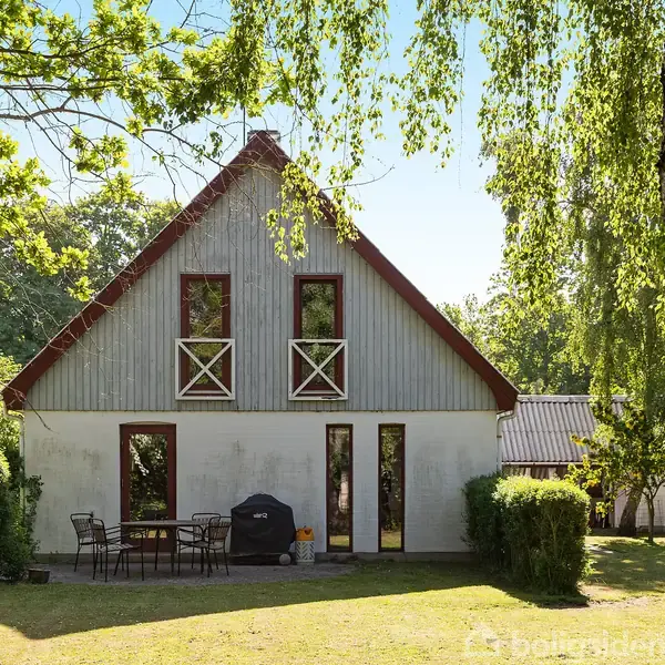 Et hvidt hus med rødt tag og to etager står i en grøn have. Et rundt bord med stole og en grill er placeret foran huset, omgivet af buske og træer.
