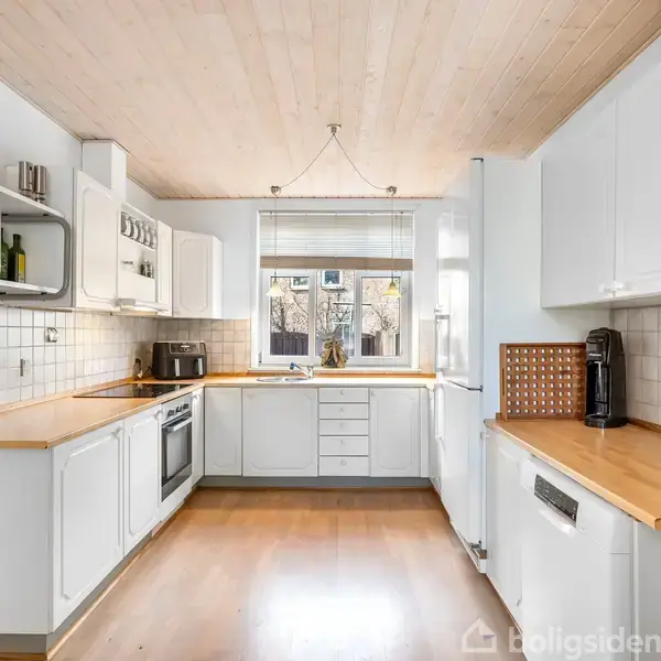 Et funktionelt køkken med hvide skabe og træbordplader. En ovn og kogeplade er integreret. Køkkenet er lyst med et vindue, der giver naturligt lys, omgivet af hvide vægge.
