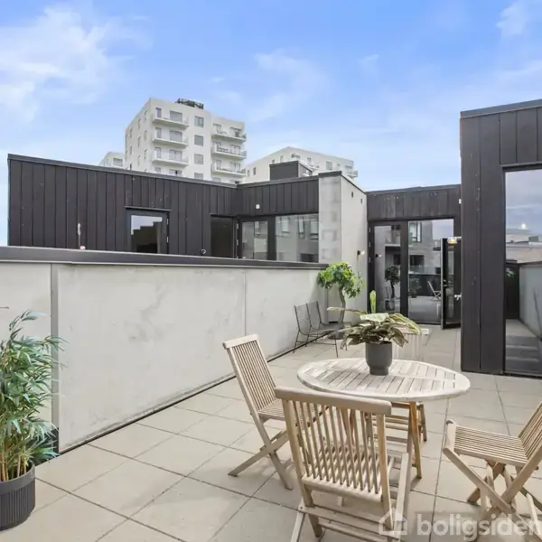 Træmøbler står på en flisebelagt tagterrasse; to stole og et bord med planter ovenpå. Baggrunden viser moderne bygninger mod en klar blå himmel.