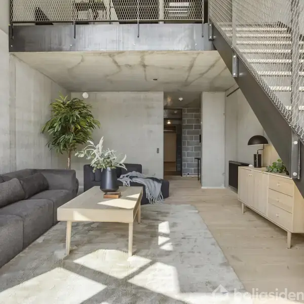 Grå sofa ved siden af et sofabord med blomster, under trappe med metalrækværk i moderne, lyst rum med betonvægge. Skab på højre side og stort plantemaleri på væggen.