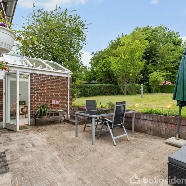 To havemøbler står stille på en terrasse med grå fliser foran en murstensbygning og glasdør til en udestue. Græsplæne og træer omkranser området.